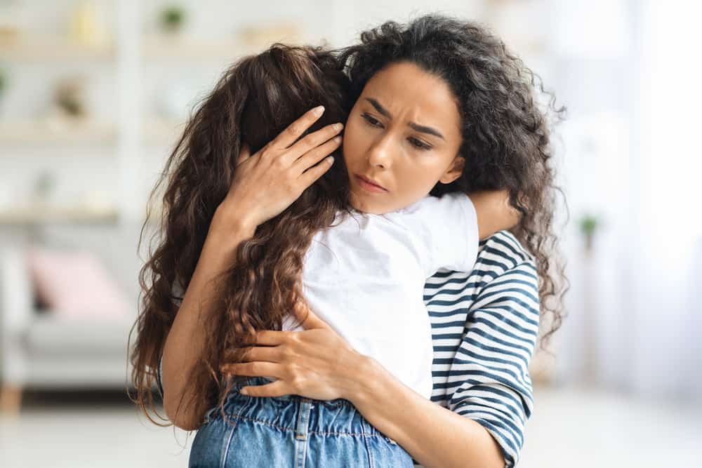 Loving mother hugging her crying little daughter, closeup, home interior. Beautiful lady comforting and embracing her upset curly girl kid. Motherhood, parenthood, mothers love concept