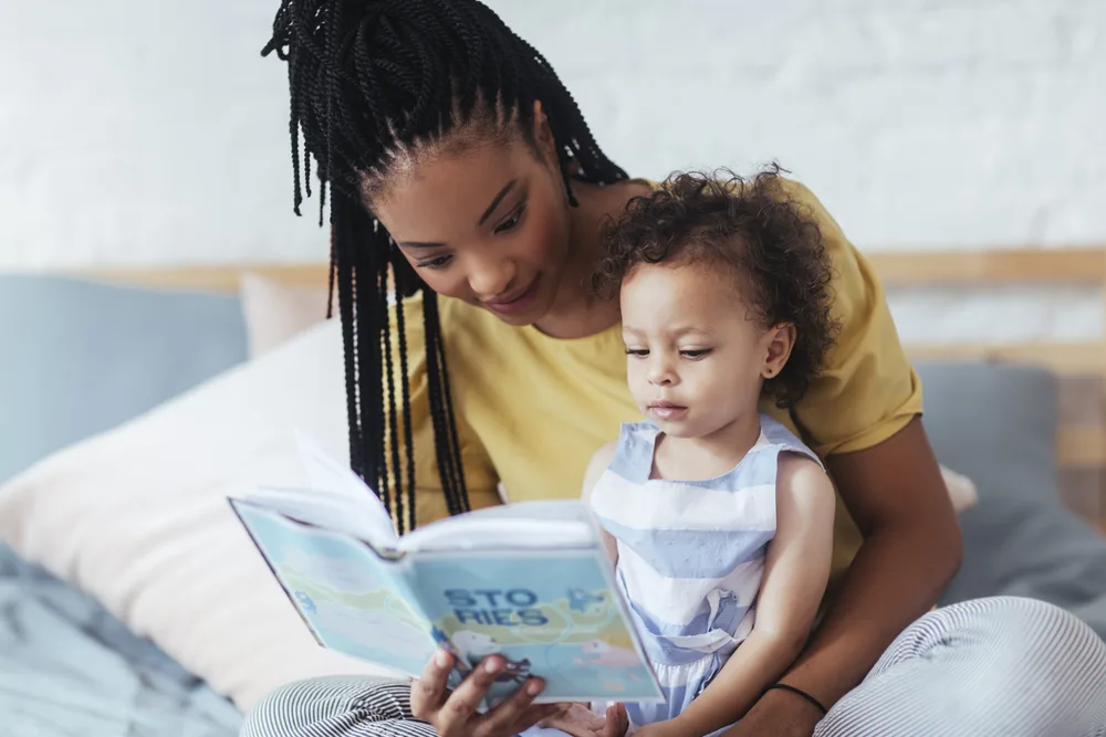 Beautiful African woman sitting on bed and reading a book to her cute baby daughter.