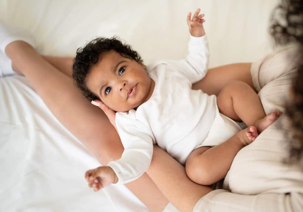 Loving young african american lady holding cute baby in her arms, enjoy tender moment on bed in light bedroom interior, top view. Family love, mother and child at home, baby care