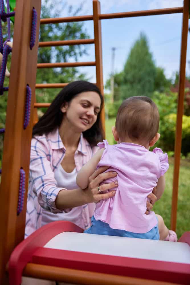 ittle baby girl with smiling mother playing on slide in playground in summer. Summer family leisure