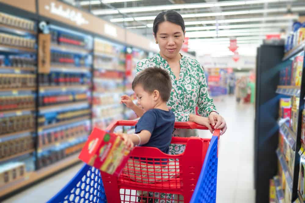 Young asian mom in green floral dress and her little multiracial boy in shopping trolley buying groceries in supermarket