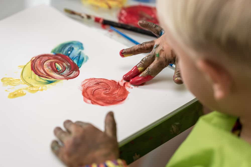 Over the shoulder view of a little boy having fun painting with fingers with colourful hand paints.