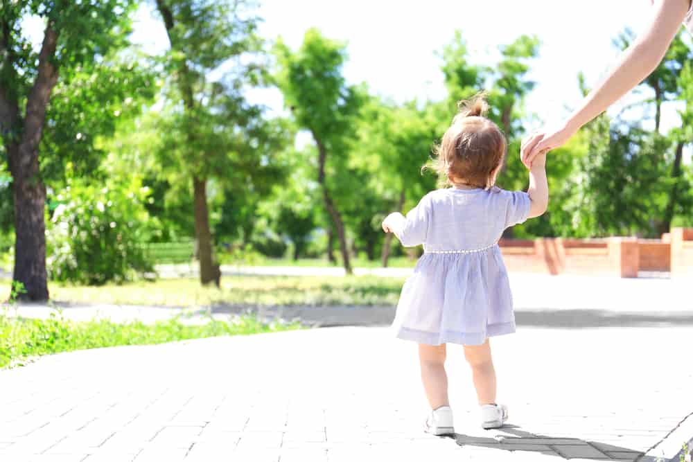 Adorable baby girl holding mother's hand while learning to walk outdoors