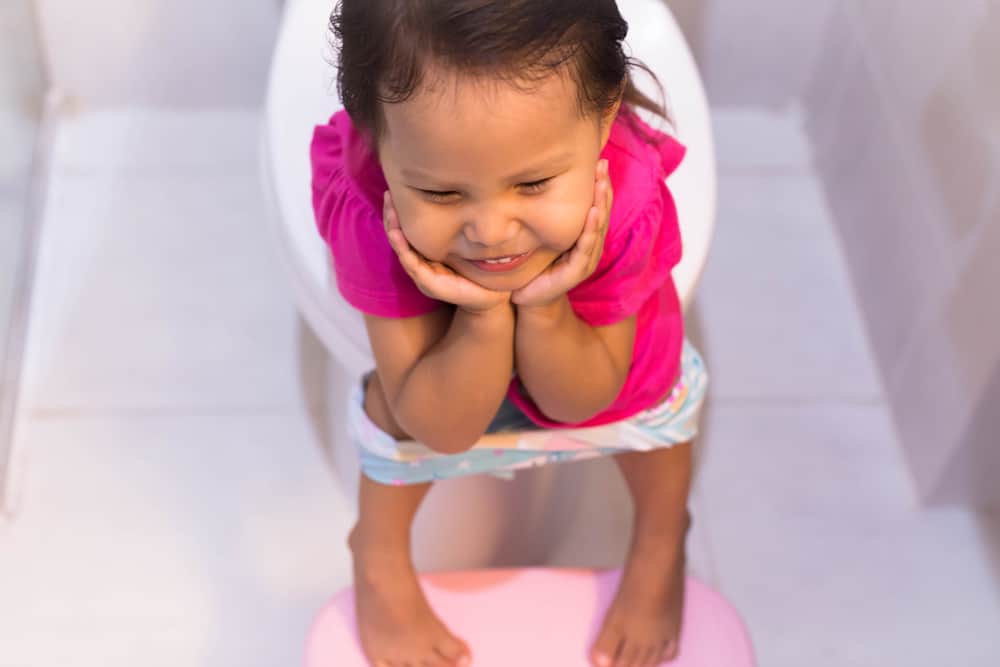 A little female child sitting on the big toilet in a white bathroom, smiling and trying to go pee.