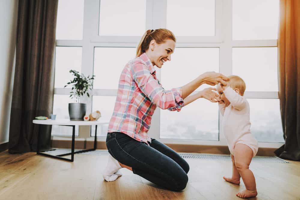 Happy Young Mother Teaching Baby Walking Indoors. Smiling Mom Wears Casual Clothes Holding Hands of Sweet Beautiful Caucasian Toddler near Large Panoramic Window in Living Room. Childhood Concept