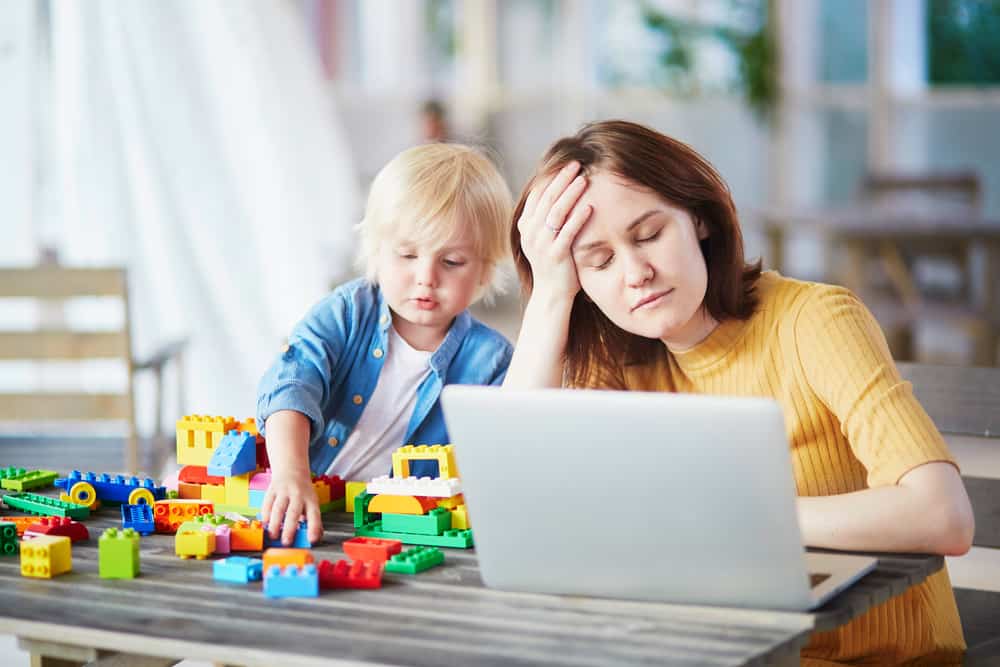 Adorable little boy playing with colorful plastic construction blocks while his mother working on her laptop. Childcare and work concept