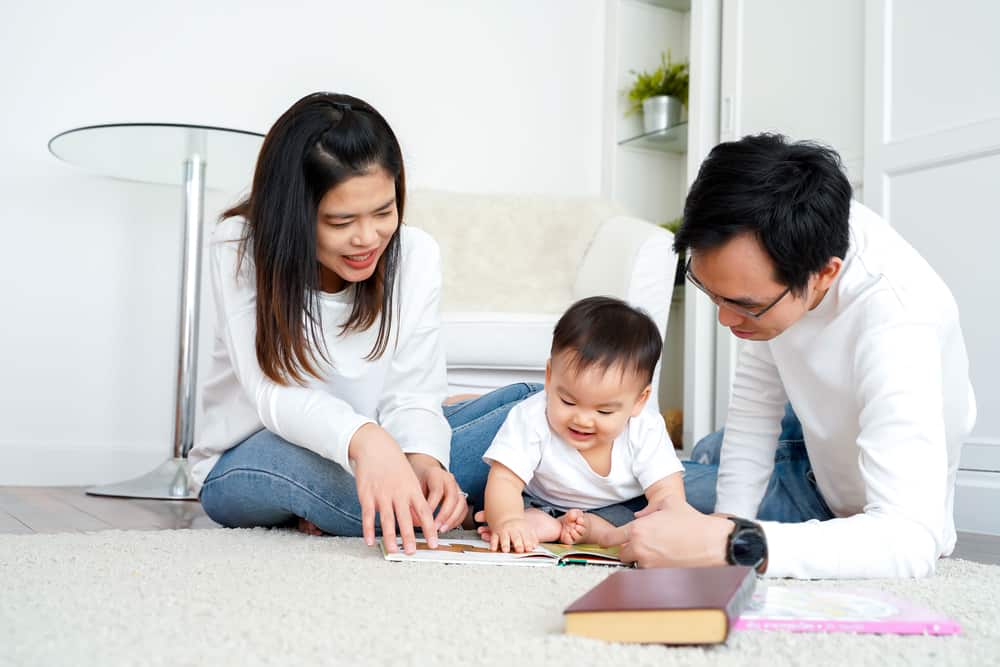 Happy modern Asian parents sitting on floor with cute infant boy and reading fairy tale while enjoying time together at home