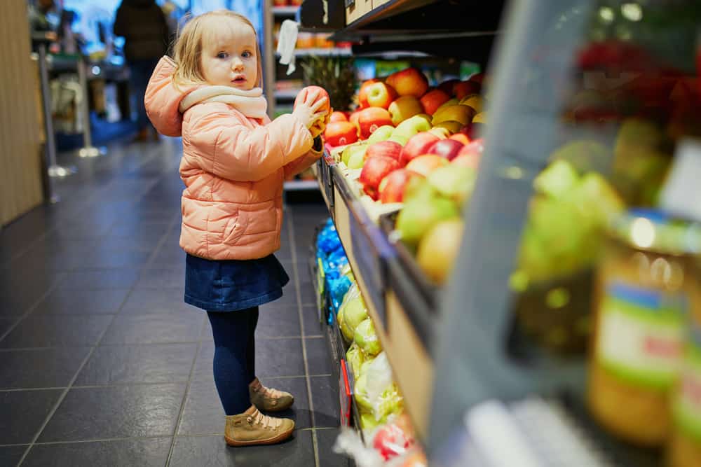 Adorable baby girl in supermarket selecting apples. Little child going shopping. Kid in large food or grocery store