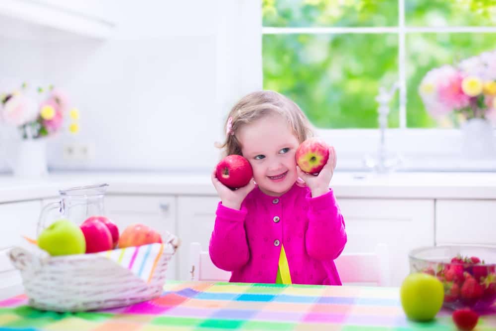Child eating breakfast. Kids eat in a white kitchen. Children having fresh fruit. Little kid playing peek a boo with apples. Cute preschooler girl with apple and strawberry on a sunny morning. Healthy nutrition for toddlers.